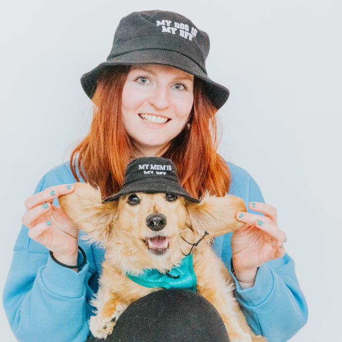 A smiling woman with red hair in a blue sweater and her dog wear matching Sassy Woof Bucket Hat Bundle - BFF hats. She lifts the dog’s ears, showing off their fun, embroidered hats against a plain white background.