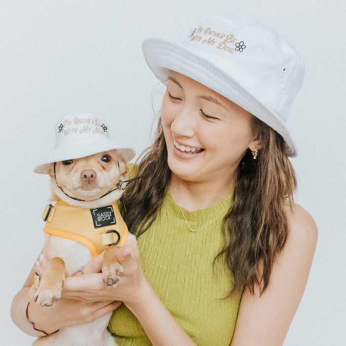 A woman in a green sleeveless top and her dog, both wearing Sassy Woof's "I'd Rather Be" Bucket Hat Bundle, pose happily together. The dog also wears a yellow harness against a plain light background.