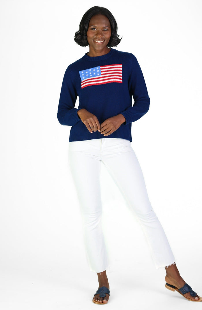 Woman wearing navy sweater with US flag design and white pants on a white background