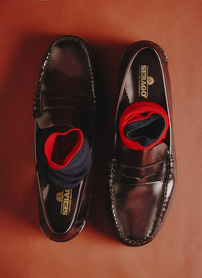 The Percival navy merino wool socks, with a contrasting red trim, styled inside a pair of dark brown leather penny loafers.