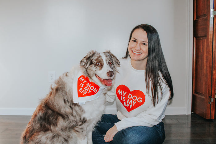 A woman in a Sassy Woof "My Dog is my BFF" (RED) sweatshirt sits smiling by her fluffy dog, who sports a matching "My Mom is my BFF" dog sweatshirt. Both sweatshirts are unisex.