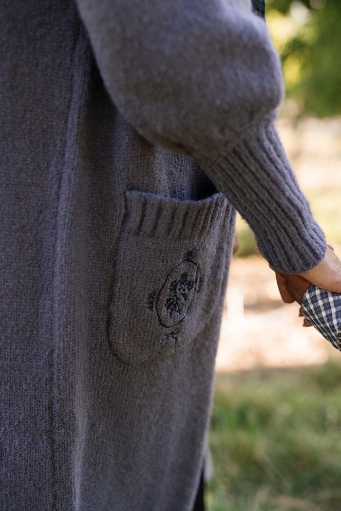 Wearing the Ivy City Co Teresa Cardigan in Dusty Blue, which features bishop sleeves and a pocket embroidered with flowers, a person holds a child’s hand outdoors. Only part of both are visible.