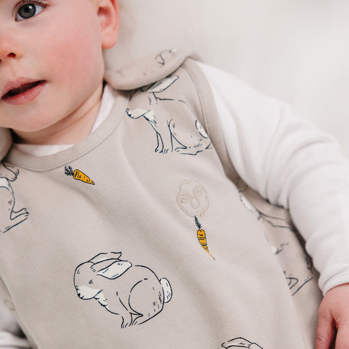Baby wearing a light gray Woolino Ultimate sleep sack with rabbit and carrot patterns on a white background