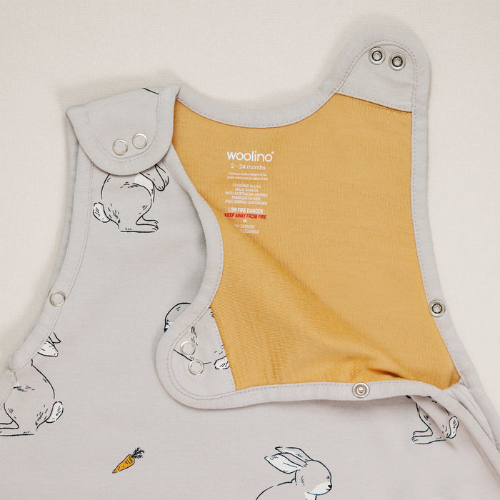 Baby sleep sack with mustard yellow inner lining and gray outer layer featuring rabbit illustrations on a light gray background.