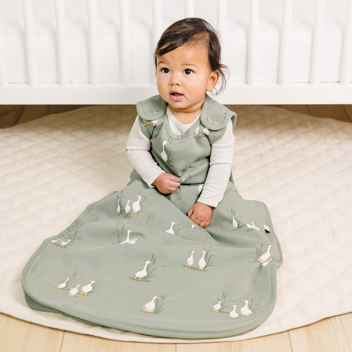 Baby sitting on a baby rug wearing a green baby bag with duck patterns