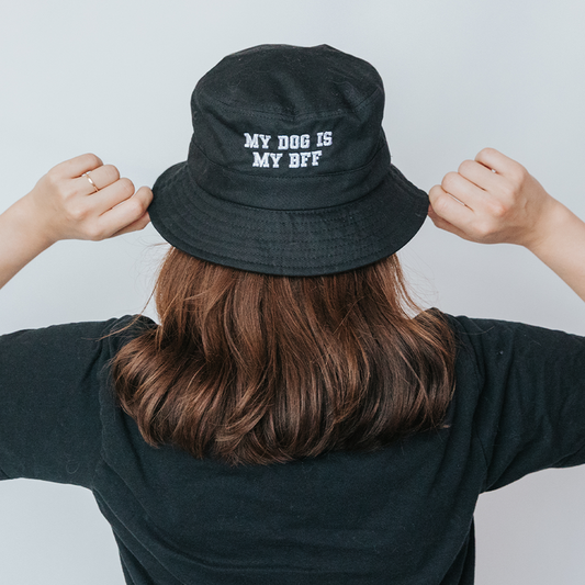A person with shoulder-length brown hair faces away from the camera, holding a Sassy Woof Bucket Hat that features "MY DOG IS MY BFF" embroidered in white.