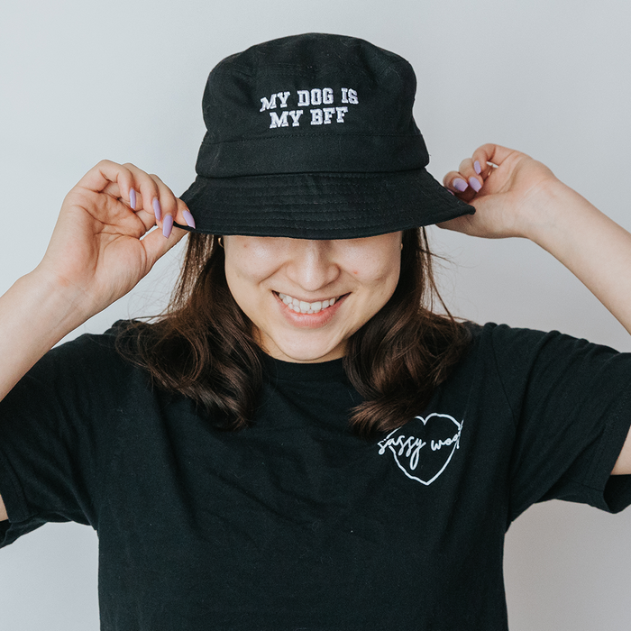 A smiling woman wears Sassy Woof's Bucket Hat Bundle - BFF, featuring a black embroidered bucket hat with "MY DOG IS MY BFF" and a matching black tee with a heart design, standing against a plain background.