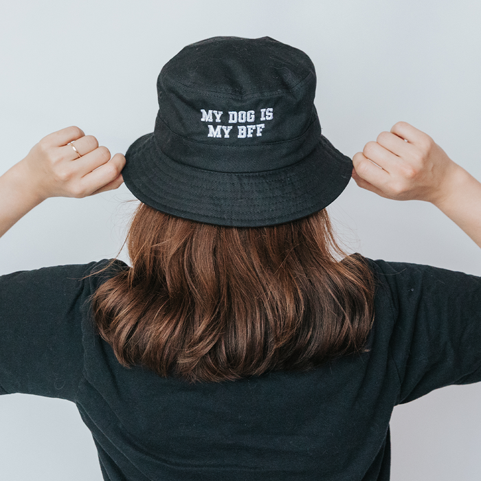 A person with brown hair, seen from behind, wears the Sassy Woof Bucket Hat Bundle - BFF, featuring “MY DOG IS MY BFF” embroidered in white. They hold the brim with both hands and wear a black shirt.