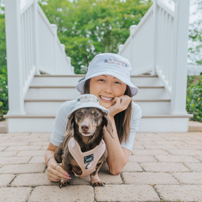 A smiling woman and a brown dachshund wear matching Sassy Woof "I'd Rather Be" Bucket Hat Bundles as they lie side by side on a brick path near outdoor steps and greenery.