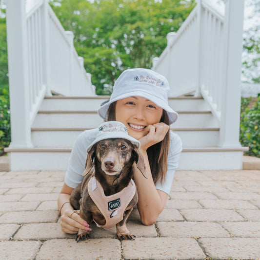 A smiling woman and a brown dachshund wear matching Sassy Woof "I'd Rather Be" Bucket Hat Bundles as they lie side by side on a brick path near outdoor steps and greenery.