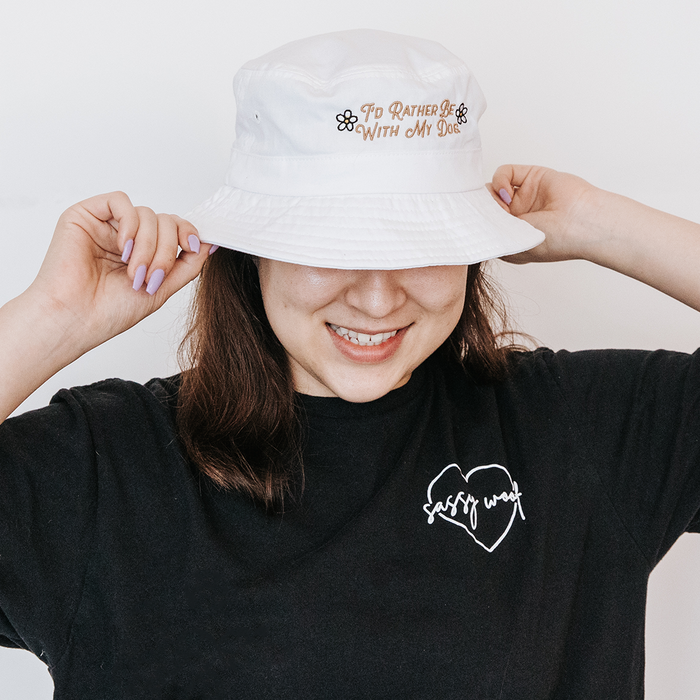 A smiling person wears the Sassy Woof Bucket Hat Bundle - I'd Rather Be, featuring an embroidered dog design and a black tee with a heart-shaped Sassy Woof logo, holding the brim of the hat with both hands.