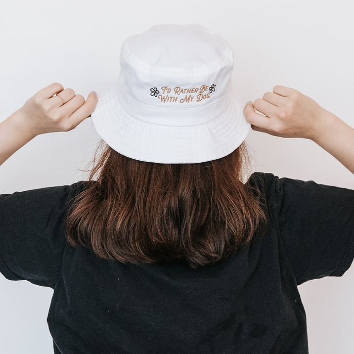 A person with long brown hair, wearing a black shirt and a Sassy Woof "Bucket Hat Bundle - I'd Rather Be" white hat with "I'd rather be with my dog" and paw prints embroidered, stands facing away.