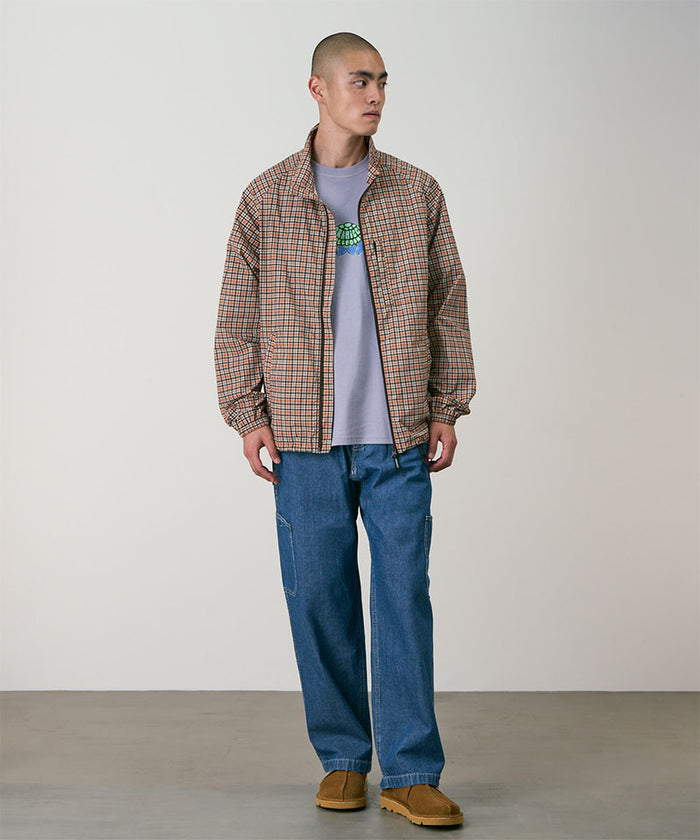 Model wearing a brick colored Gramicci Checkered Shell Jacket and blue pants on a light-tone background.