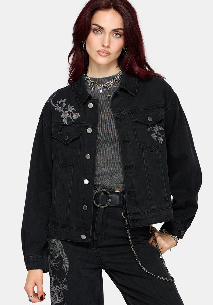 A model with dark red hair wearing a black denim jacket with gray floral embroidery on the chest, layered over a gray textured top and black jeans.