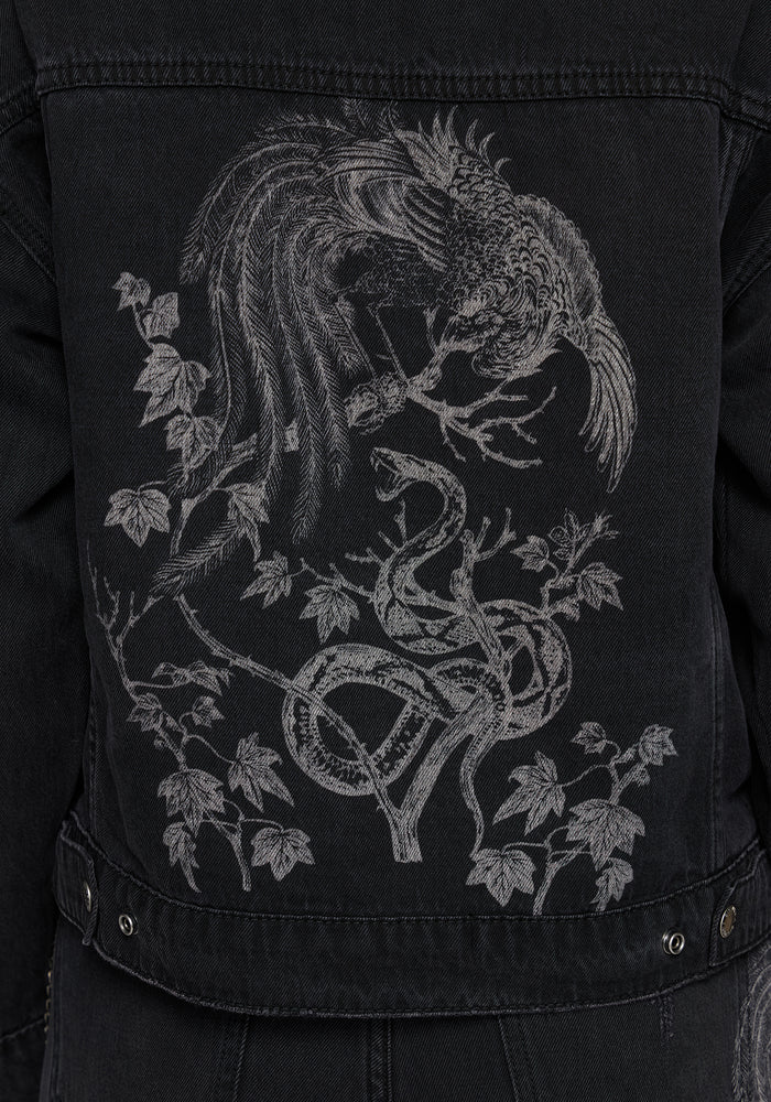 Close-up of the back of a black denim jacket featuring a large white embroidered graphic of a bird perched on a tree with a snake and leaves.