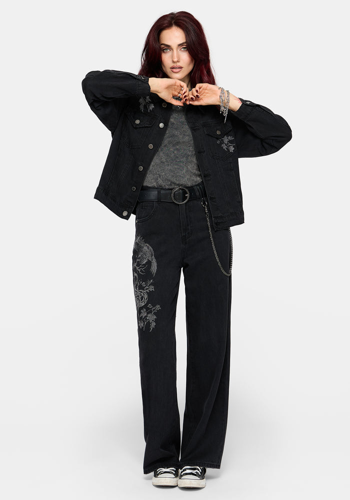 A model wearing a black denim jacket with floral embroidery, a gray knit top, black wide-leg jeans with matching embroidery, a black belt, and black sneakers, posed against a light gray studio background.