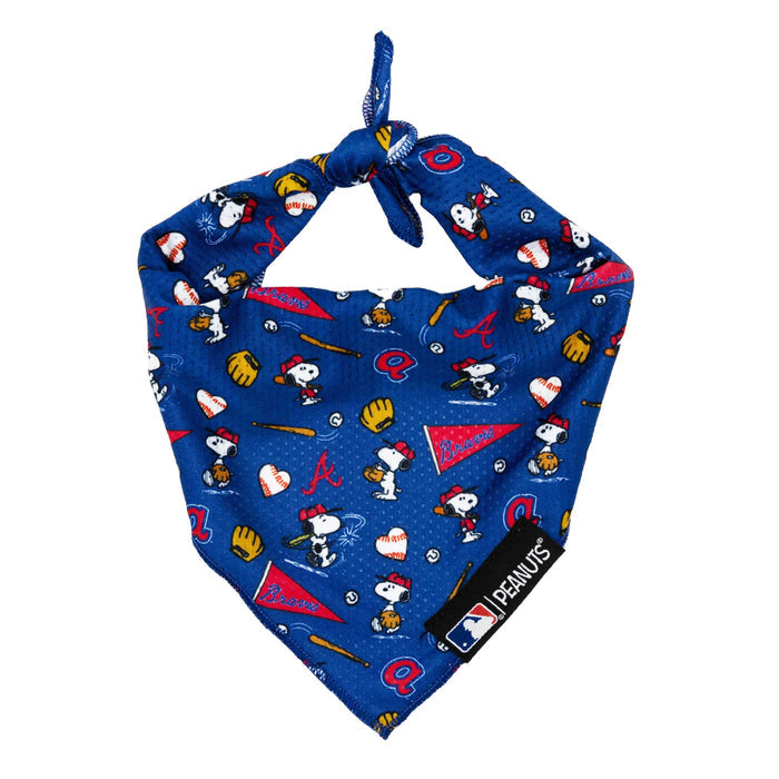 The Sassy Woof Dog Bandana - MLB™ x Peanuts™ Atlanta Braves™ is a blue bandana with a top knot, showcasing Snoopy and Woodstock playing baseball among Braves logos. A black tag at the bottom displays the MLB and Peanuts logos.