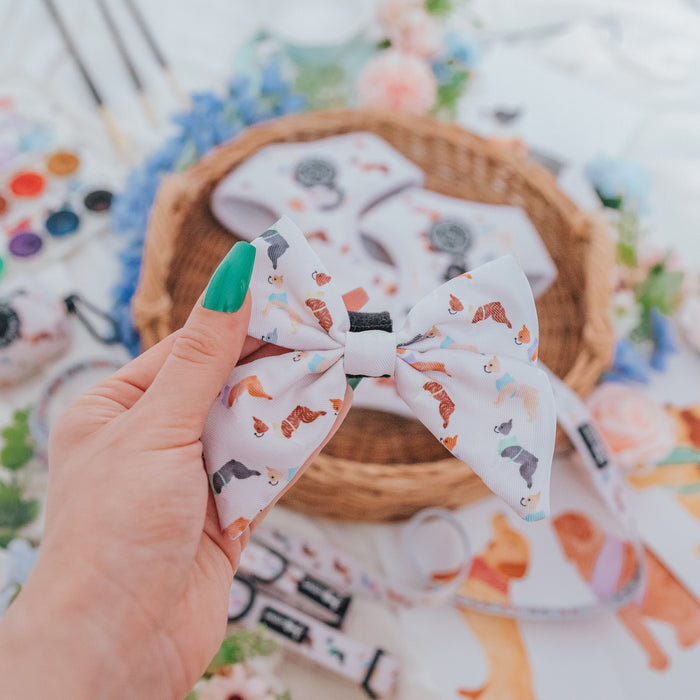 A hand with green-painted nails holds the Sassy Woof Dog Sailor Bow - Bold Boston Terriers in front of a wicker basket filled with matching harness and collar pet accessories, surrounded by colorful art supplies and dog-themed items.