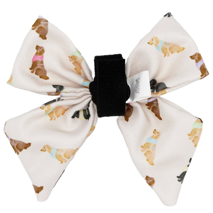 The Sassy Woof Dog Sailor Bow - Loyal Labradors is a white fabric bow tie with a black center, featuring illustrated dogs in colorful sweaters.