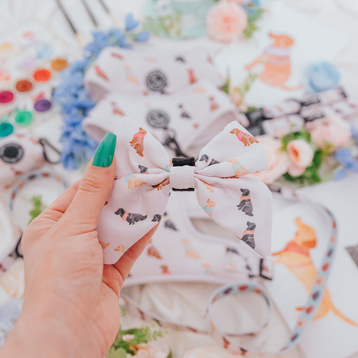 A hand with green nail polish holds the Sassy Woof Dog Sailor Bow - Loyal Labradors, featuring colorful dog illustrations, surrounded by matching accessories like an adjustable harness, flowers, and art supplies in the background.