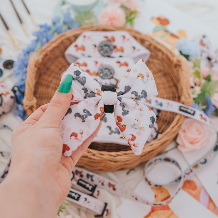 A hand with green-painted nails holds the Sassy Woof Dog Sailor Bow - Spunky Shih Tzus, featuring a colorful dog pattern, in front of a wicker basket filled with matching accessories and surrounded by flowers.
