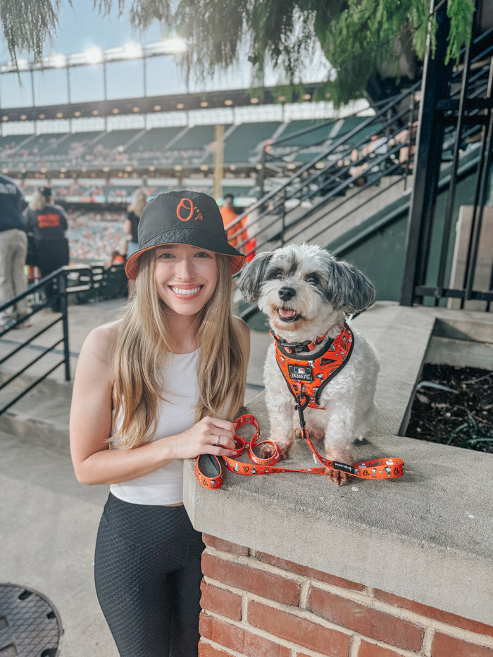 A smiling woman in an Orioles hat poses with her black-and-white dog, who’s wearing a Sassy Woof Dog Adjustable Harness - MLB™ x Peanuts™ Baltimore Orioles™, at a baseball stadium filled with fans.