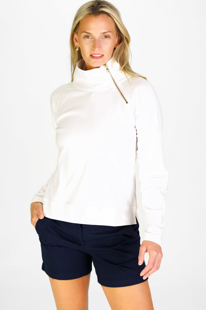 Woman wearing a white long-sleeve top with a gold asymmetrical zipper and navy shorts on a white background