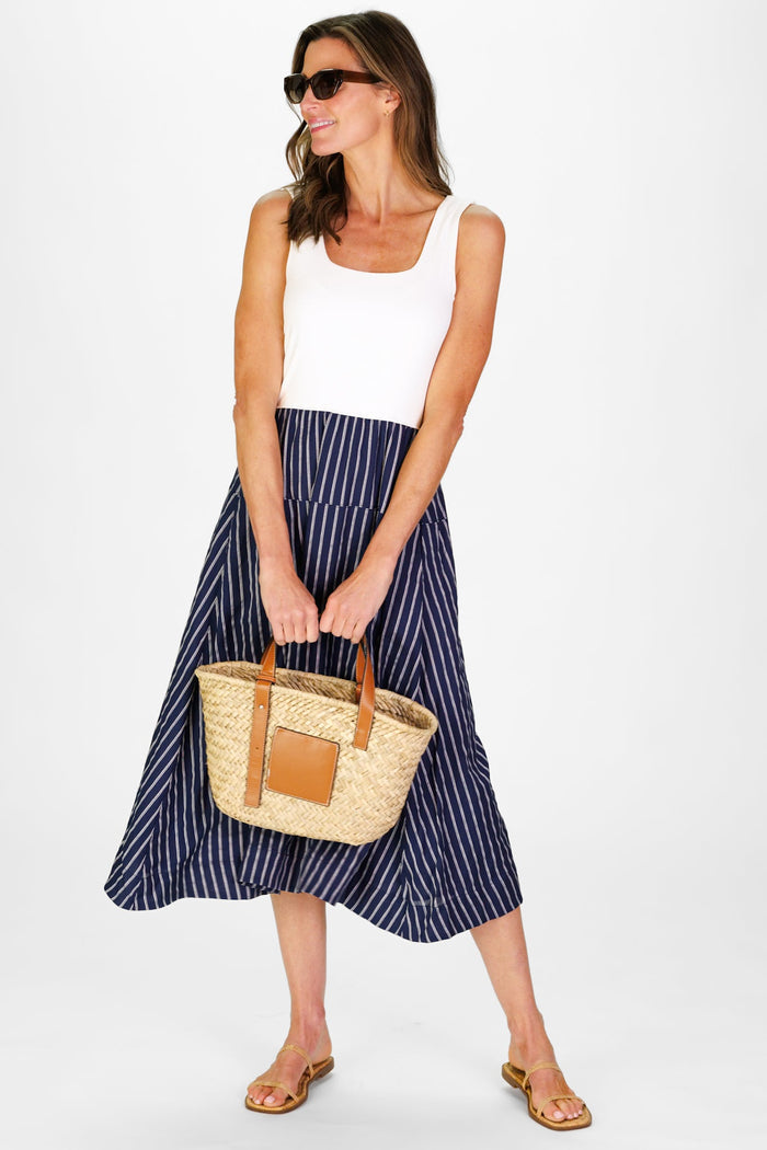 Model In the Duffield Lane Jada Dress, a midi length dress with a white cotton bodice and a linen navy skirt with white stripes, holding a woven bag
