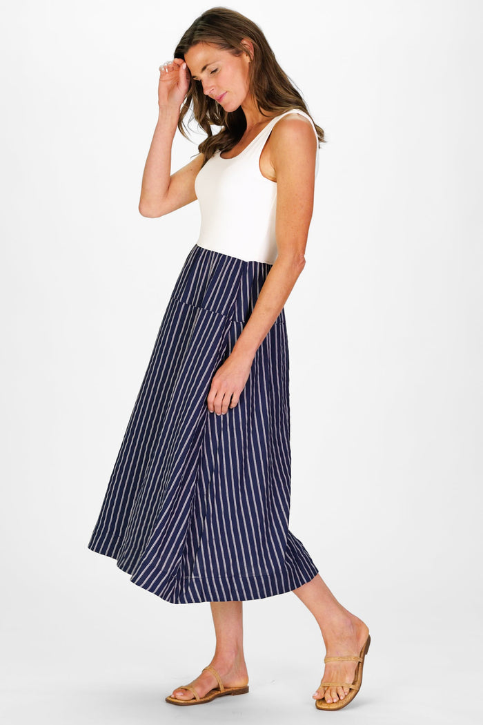 Model In the Duffield Lane Jada Dress, a midi length dress with a white cotton bodice and a linen navy skirt with white stripes, holding a woven bag