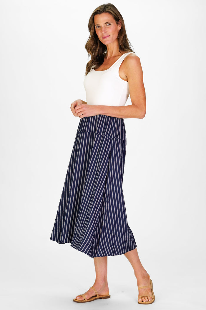 Model In the Duffield Lane Jada Dress, a midi length dress with a white cotton bodice and a linen navy skirt with white stripes, holding a woven bag