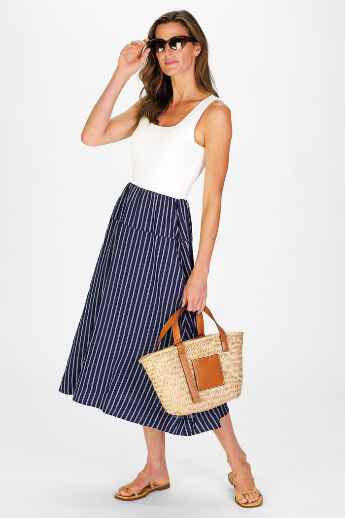 Model In the Duffield Lane Jada Dress, a midi length dress with a white cotton bodice and a linen navy skirt with white stripes, holding a woven bag