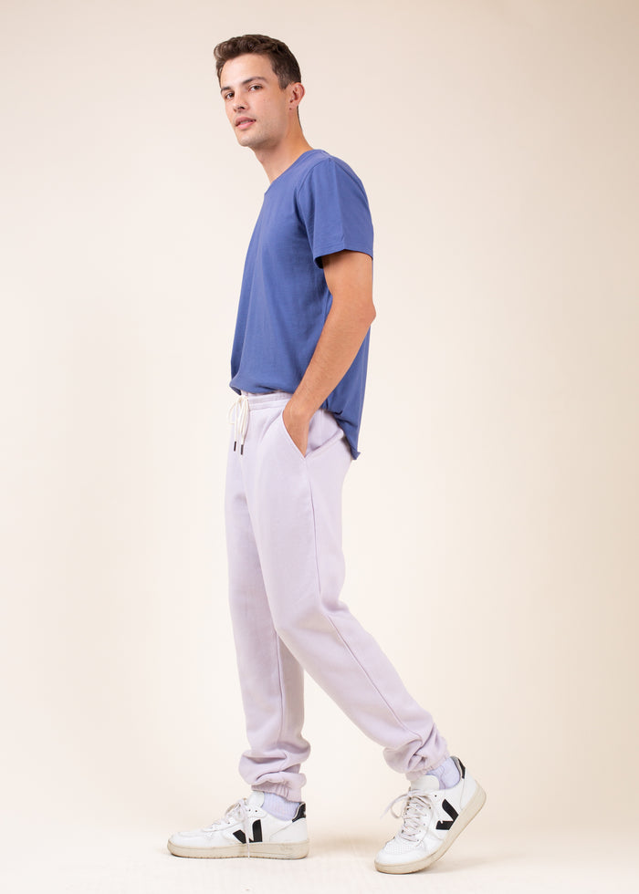 Man wearing a blue shirt, light purple sweatpants, and white sneakers, frorm the side,  on a beige background