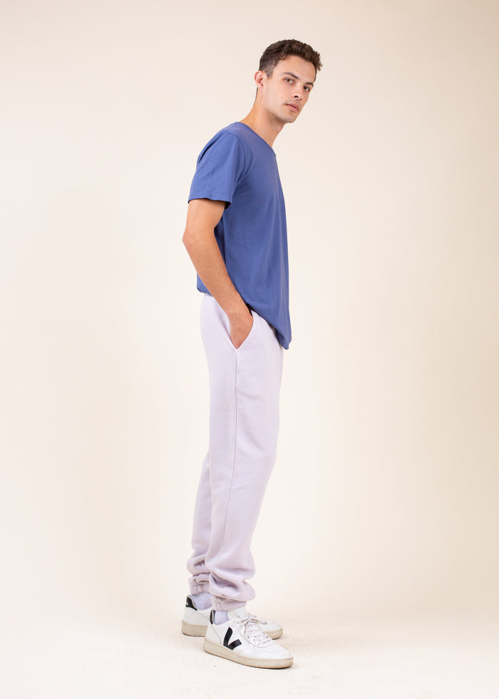 Man wearing a blue t-shirt, light purple sweatpants, and white sneakers, viewed from thje side, on a beige background