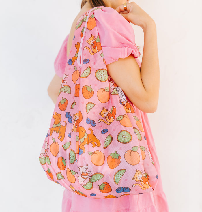 A person in a pink dress holds the Sassy Woof Fruity Reusable Bag, which features playful cat illustrations, fruit slices, and berries, set against a plain white background.