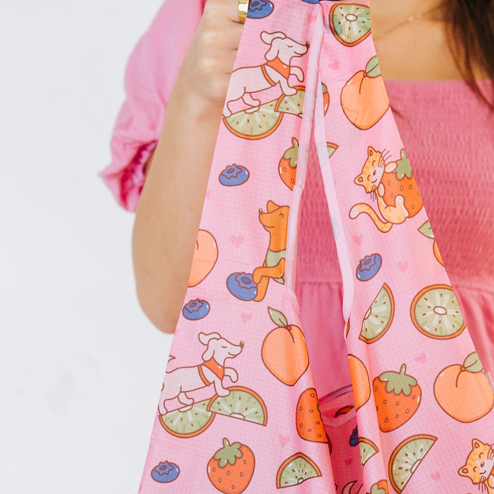 A person in a pink top holds the Sassy Woof Fruity Reusable Bag, featuring playful dog, cat, fruit, and heart designs.