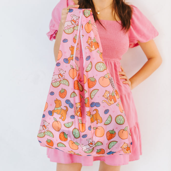 A woman in a pink dress holds the Sassy Woof Fruity Reusable Bag, a pink foldable shopping bag featuring colorful cat, dog, fruit, and vegetable illustrations, against a plain white background.