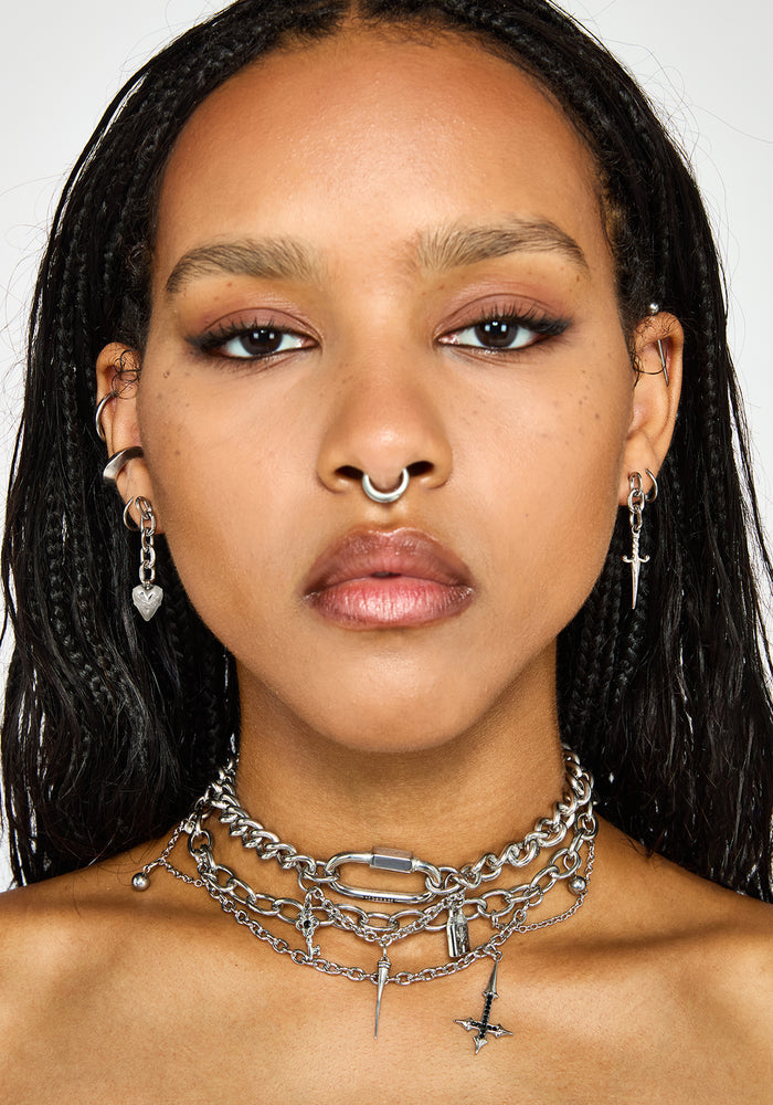 A close-up portrait of a model wearing a layered silver chain choker necklace with barbell and cross charms, along with matching silver hoop and dangle earrings.