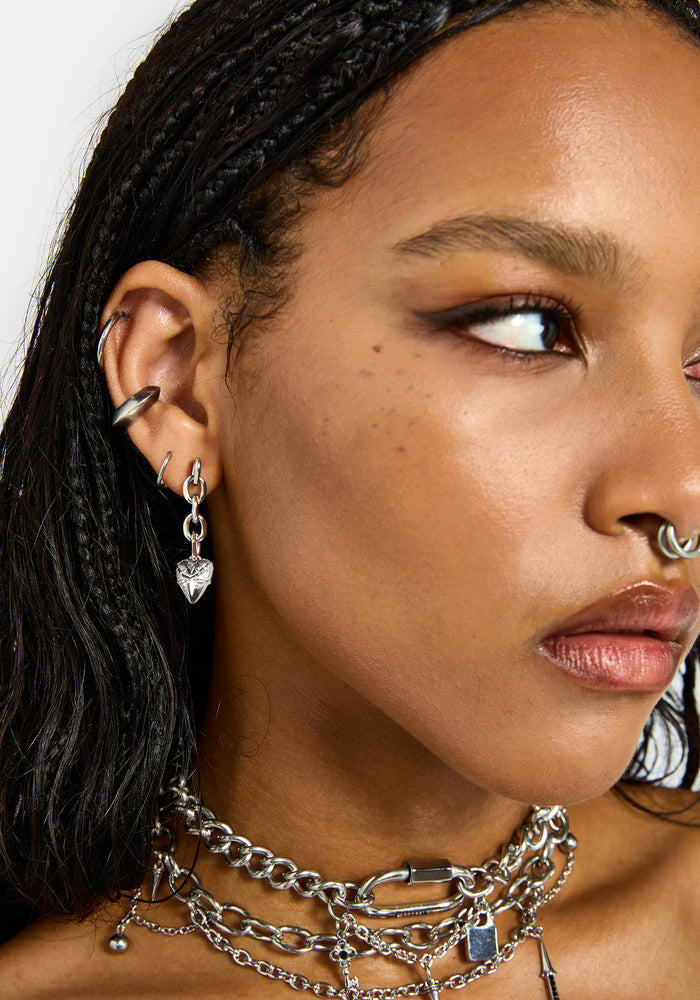 Close-up of a model wearing a silver dangling heart earring, along with multiple silver chain necklaces, hoop earrings, and a nose ring.
