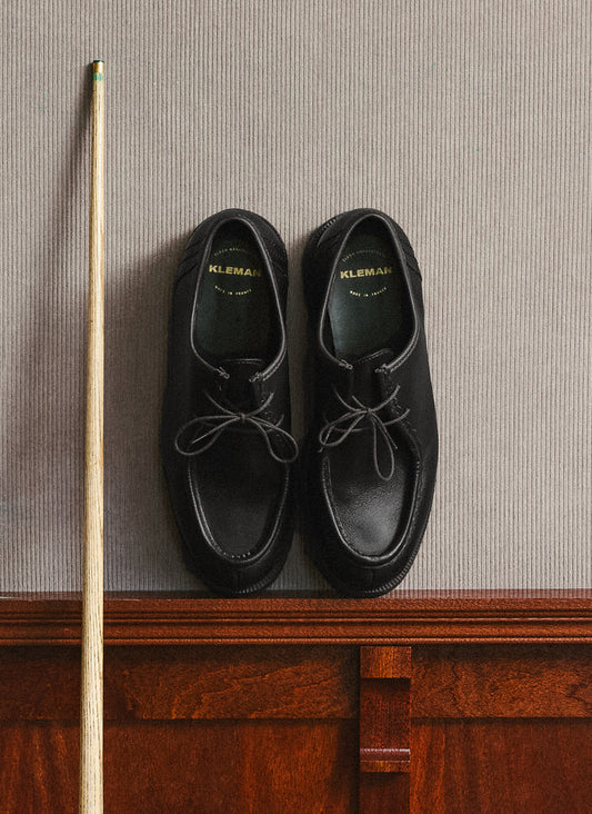 A pair of black Percival Kleman Padror shoes placed against a textured wall next to a cue stick.