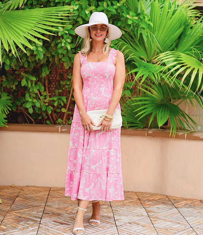 Woman in a pink dress and white hat standing in front of green plants.