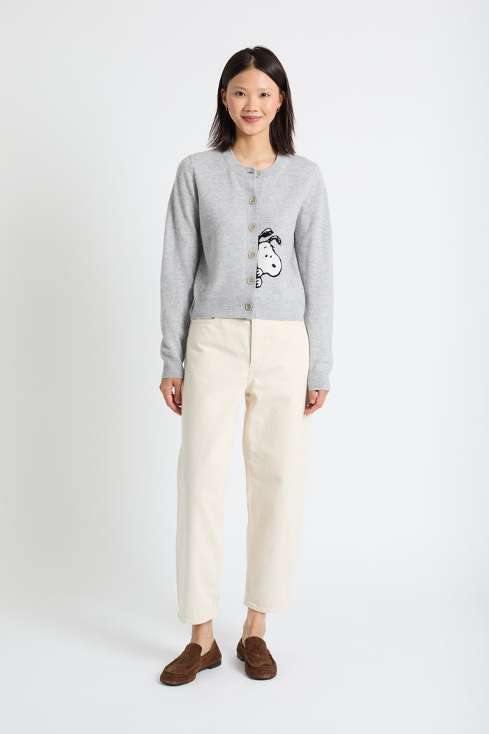 Woman wearing a grey cardigan with a graphic design, white pants, and brown shoes on a white background