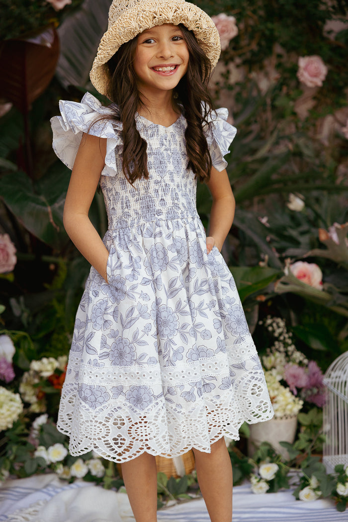 A young girl in the Ivy City Co Mini Hattie Dress in Blue Floral stands outdoors, smiling with her hands in her pockets, surrounded by lush greenery and blooming flowers.