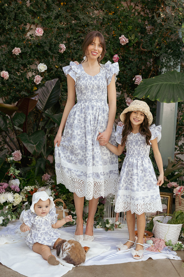 A woman and two young girls wear matching Ivy City Co Mini Hattie Dresses in Blue Floral with lace hems, posing outdoors among flowers and greenery. The older girl smiles in her Girls Mini Hattie Dress, wearing a hat by the picnic blanket and guinea pig.