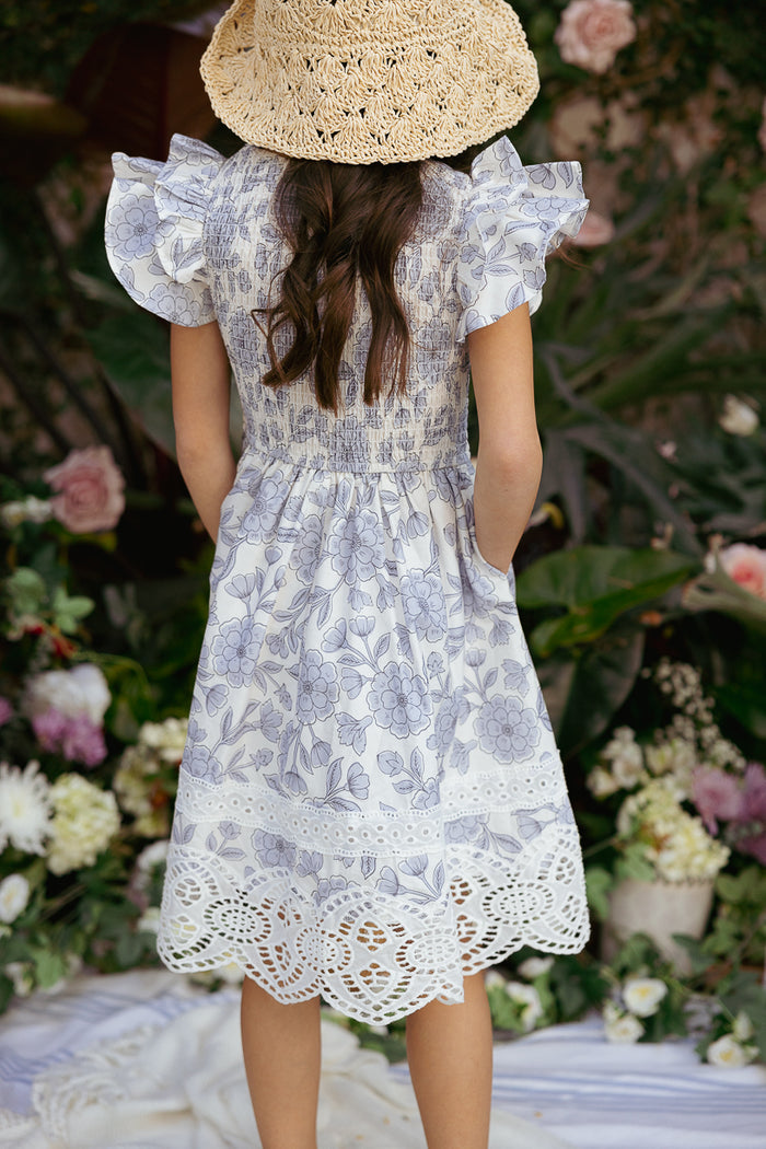 A young girl wearing the Ivy City Co Mini Hattie Dress in Blue Floral and a wide-brimmed straw hat stands outdoors, facing away, surrounded by flowers and greenery.