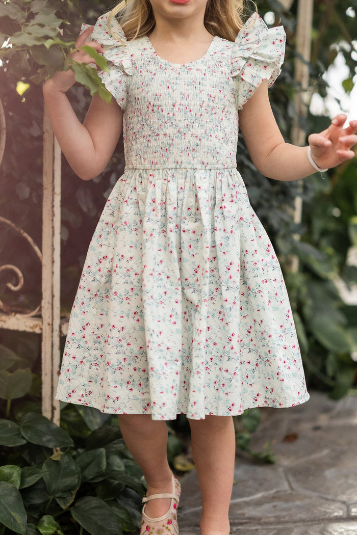 A young girl in the Ivy City Co Mini Hattie Dress in Pansy Trellis, with ruffled sleeves, stands among green plants with arms raised and face out of frame—perfect for a charming girls’ cotton floral dress.