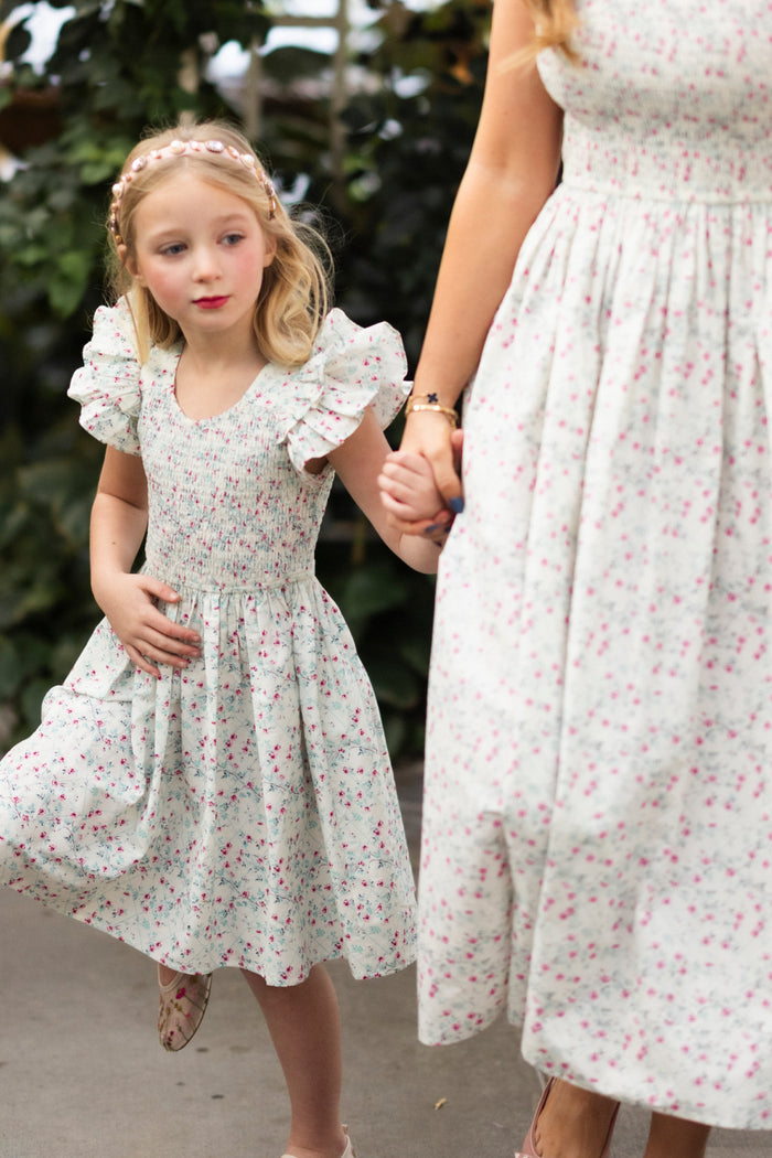 A young girl wears the Ivy City Co Mini Hattie Dress in Pansy Trellis, holding hands with an adult in a matching floral dress as they stand on a paved path surrounded by greenery. The girl glances to the side.