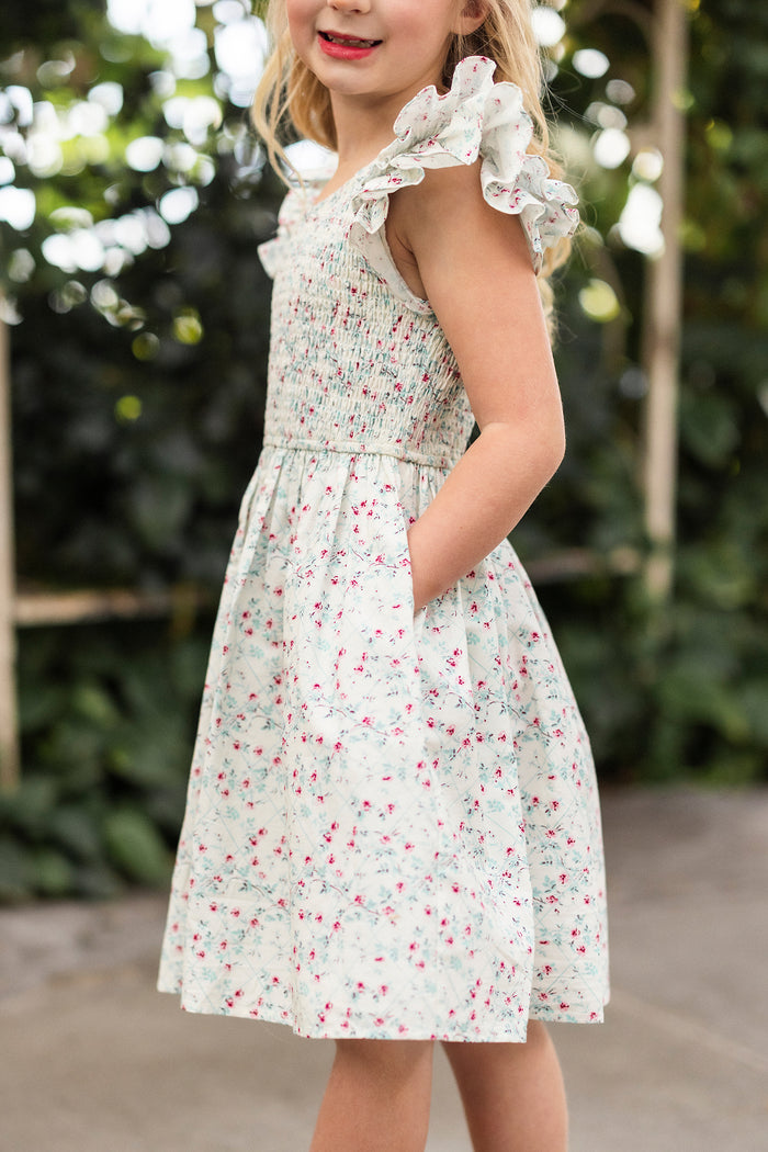 A young girl in the Ivy City Co Mini Hattie Dress in Pansy Trellis, featuring ruffled sleeves, stands smiling outdoors with her hand in her pocket. The background shows blurred green foliage.