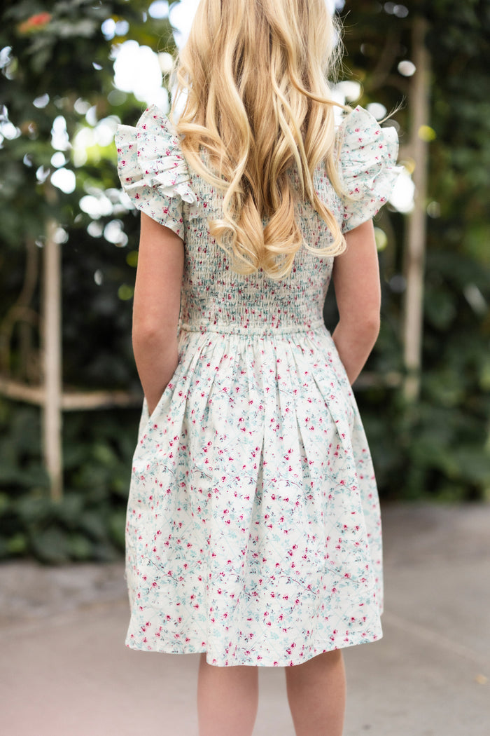 A young girl with long blonde hair stands outdoors, facing away. She wears the Ivy City Co Mini Hattie Dress in Pansy Trellis, featuring puffed short sleeves and a spring floral print. Soft green foliage is blurred in the background.