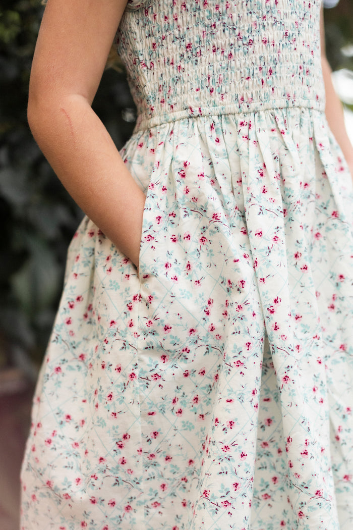 A child stands outdoors in the Ivy City Co Mini Hattie Dress in Pansy Trellis, featuring a sleeveless cut, smocked bodice, and delicate red and blue floral patterns—a charming spring floral dress.