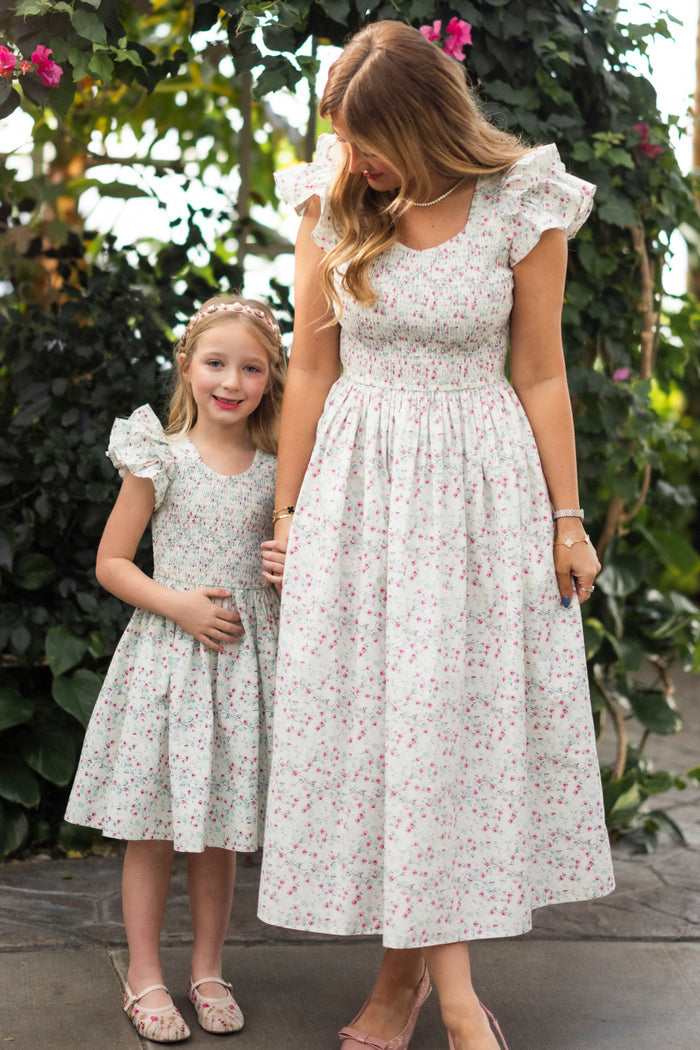A woman and young girl hold hands, smiling in matching Ivy City Co Mini Hattie Dress in Pansy Trellis. Surrounded by green foliage and flowers, their charming cotton dresses brighten the outdoor garden scene.
